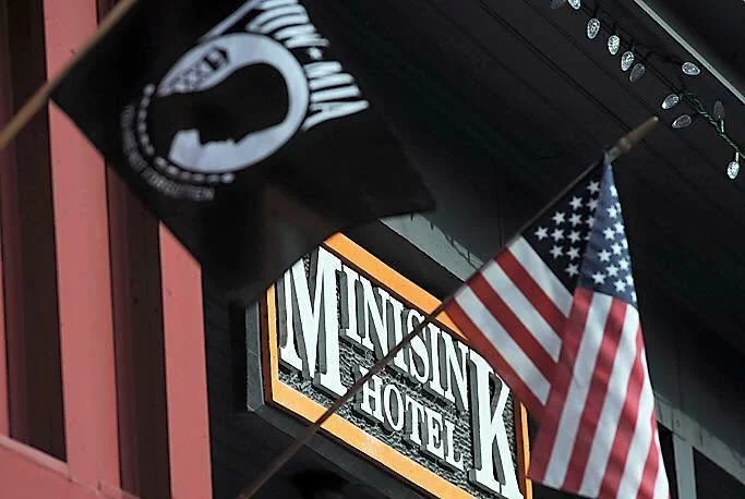 The carved Minisink Hotel sign under the porch roof, with a POW/MIA flag and an American flag against the red clapboard